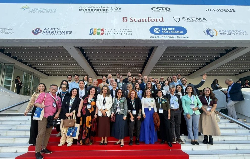 Grupo de participantes de uma conferência internacional posa para fotografia em uma escadaria com tapete vermelho. Ao fundo, aparecem logotipos de instituições e empresas ligadas à inovação, como Stanford, SKEMA e Sophia Antipolis